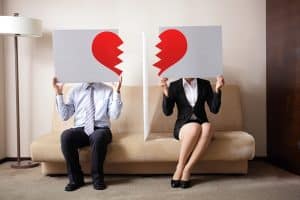 Two people sit on a beige couch, each holding a large sign with half of a broken red heart, facing forward. The person on the left wears a suit and tie, while the one on the right opts for a black blazer and skirt. Theyre contemplating an annulment in Colorado, with a floor lamp softly illuminating the scene. divorce attorney colorado springs