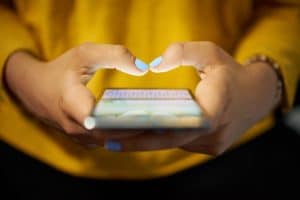 Close-up of a persons hands with blue nail polish holding a smartphone, scrolling through social media. They type intently on the device while wearing a yellow top, highlighting the risk of distraction. The background is softly blurred. divorce attorney colorado springs