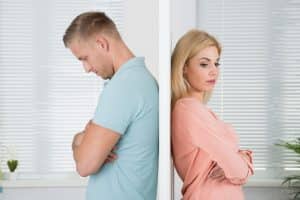 A man and woman stand back-to-back, separated by a white wall, embodying Colorado divorce tricks. The man, in a light blue shirt, looks down with arms crossed. The woman, in a peach blouse, looks away, also with crossed arms. Both appear upset. divorce attorney colorado springs