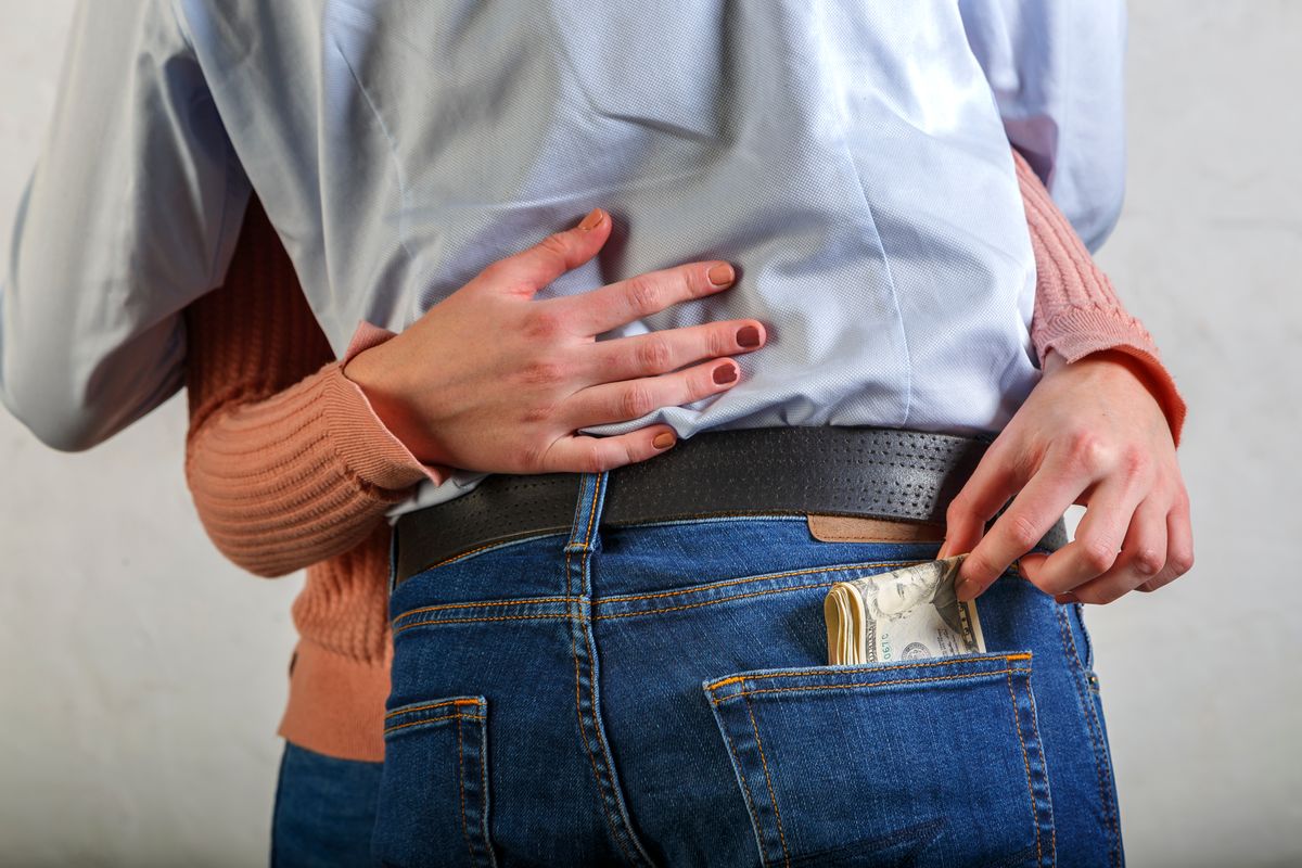 A person in a pink sweater hugs another wearing a light blue shirt and jeans. While the embrace seems affectionate, a hand discreetly takes a roll of cash from the jean pocket, an act that could raise serious charges against your spouse if discovered. divorce attorney colorado springs