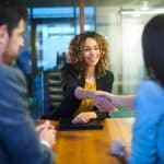 In a meeting room, a woman in a yellow top warmly shakes hands with a woman in a blue jacket. Nearby, a man in a suit observes. As sunlight filters through the glass wall, they discuss questions to ask a family attorney, ensuring clarity and confidence moving forward. divorce attorney colorado springs