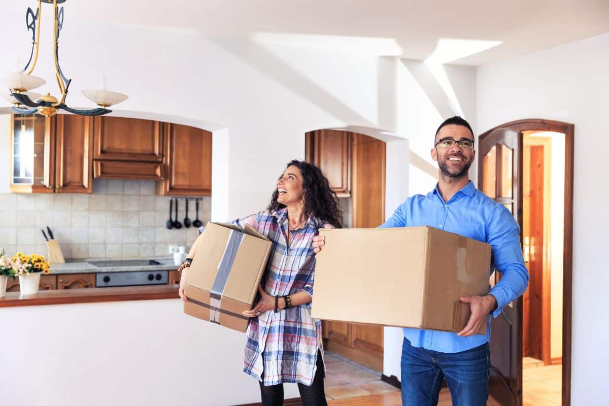 A smiling couple, in a joyful celebration of their domestic partnership, carries cardboard boxes into a bright, unfurnished kitchen. The space features wooden cabinets, a stove, and a tiled backsplash. They look excited and happy in their new home. divorce attorney colorado springs